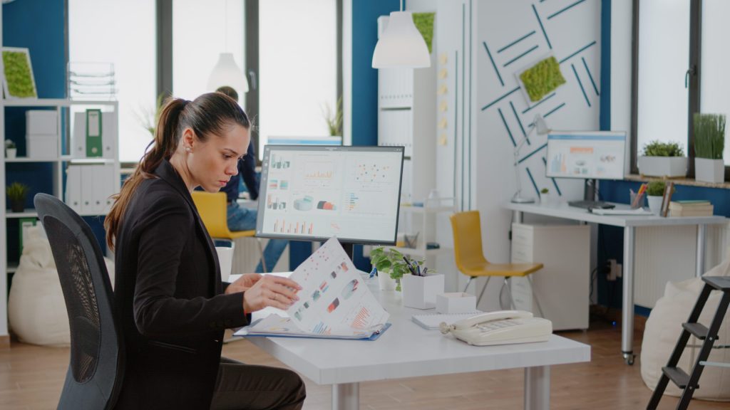 Geschäftsfrau arbeitet mit dem Computer und Datendiagrammen auf Papier, nutzt Technologie für Entwicklung und Management. Unternehmerin plant eine Marketingstrategie für ein erfolgreiches Finanzunternehmen.