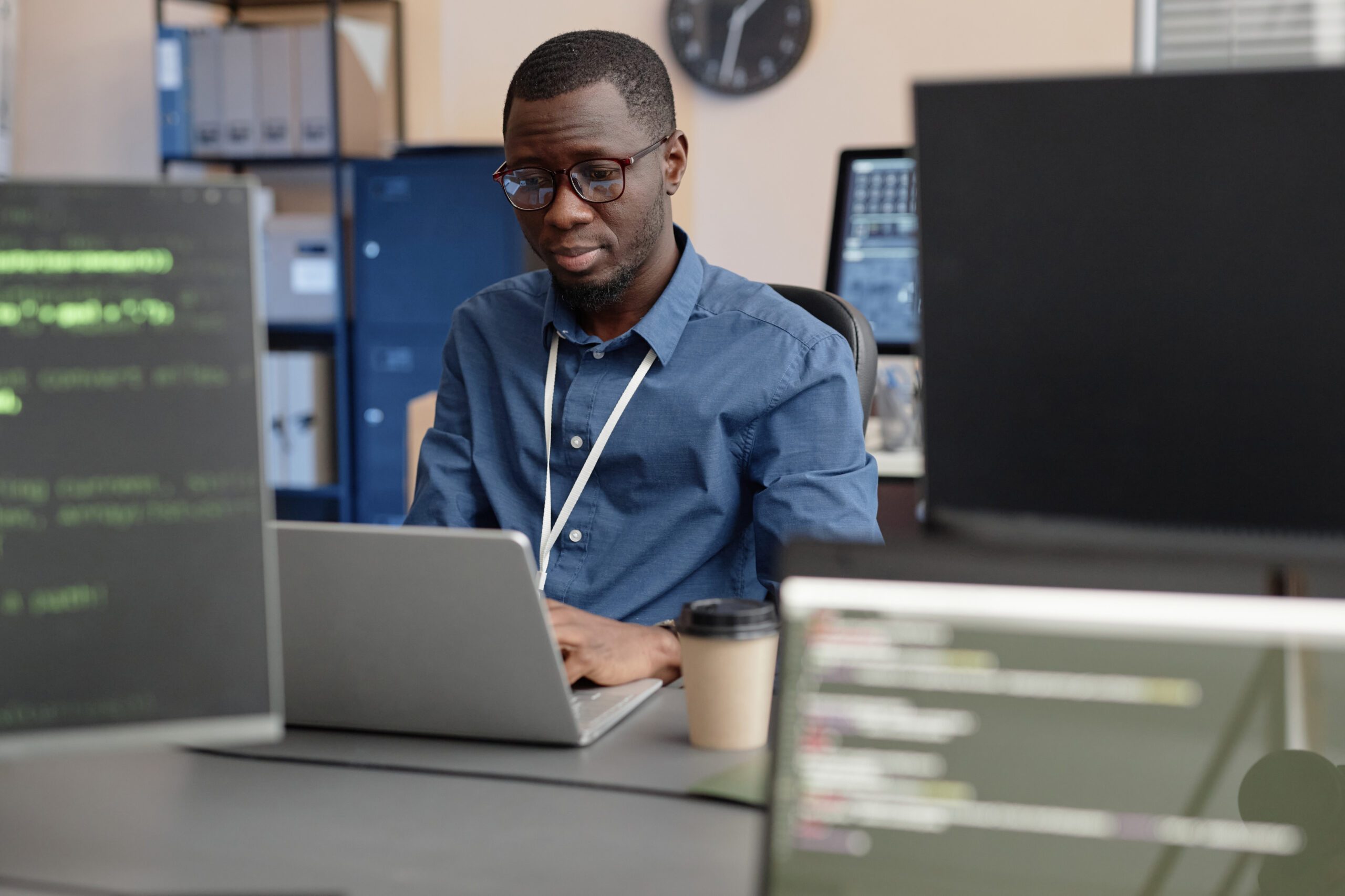 Halbtotalaufnahme eines konzentrierten afroamerikanischen Programmierers mit Brille, der auf einem Laptop tippt, während er am Schreibtisch sitzt. Der Blick geht durch verschwommene Computerbildschirme im Vordergrund.