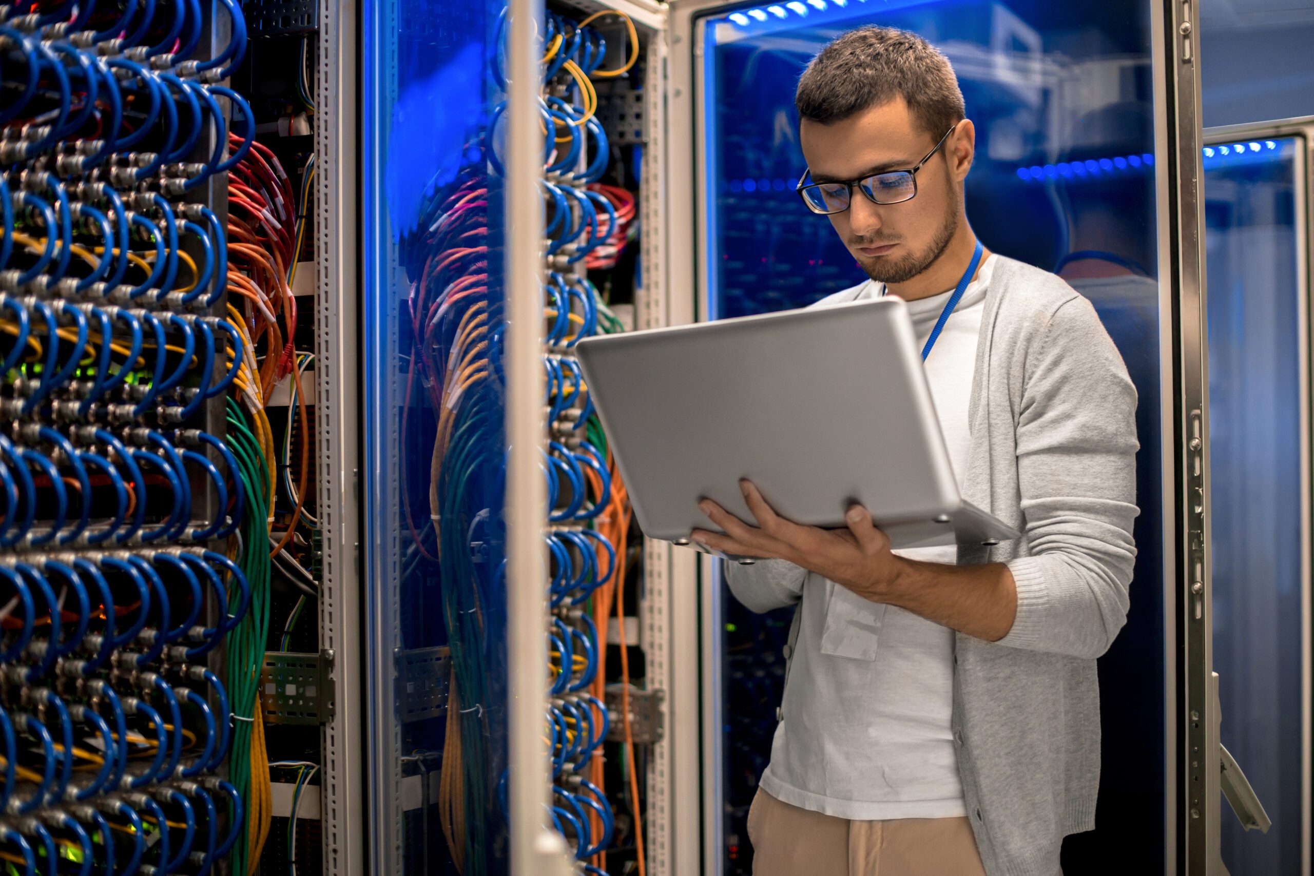 Porträt eines modernen, jungen Mannes, der einen Laptop hält und im Serverraum steht, während er mit einem Supercomputer in blauem Licht arbeitet.